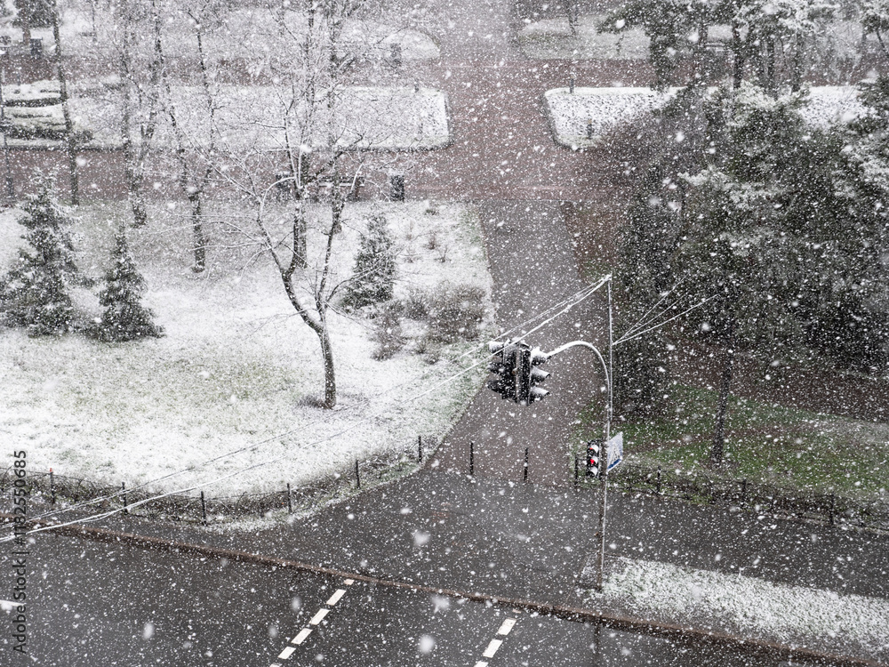View from the window over heavy fluffy snow falling over the pedestrian crossing with red traffic light and urban park with a lonely person walking. Snowy winter day.