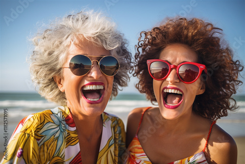 Two older women laughing hysterically and smiling on a beach. The girls are wearing sunglasses. One has gray hair and the other brown. Ladies enjoying their vacation.