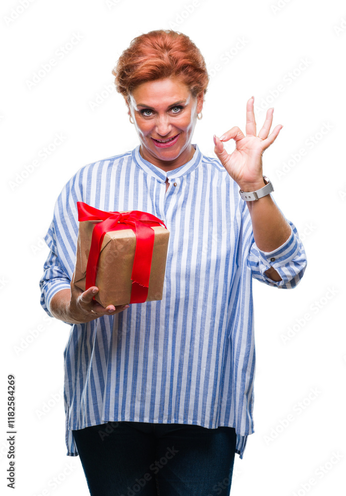 Senior caucasian woman holding gift over isolated background doing ok sign with fingers, excellent symbol Senior caucasian woman holding gift over isolated background doing ok sign with fingers, excellent symbol