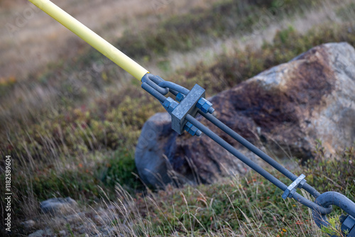 A guy wire secured to the ground from a distribution utility pole. The galvanized wire is wrapped in a yellow insulator covering. The anchor and tensioned stability cables are connected to a hook. 
