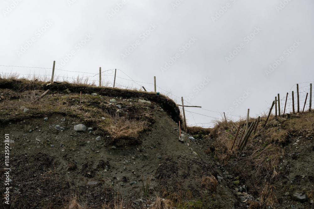 The edge of a cliff with a top layer of hay and grass. The rich topsoil is naturally moving downward. The ground has eroded and is undermined below the sod section of the ground. The edge wire fence.