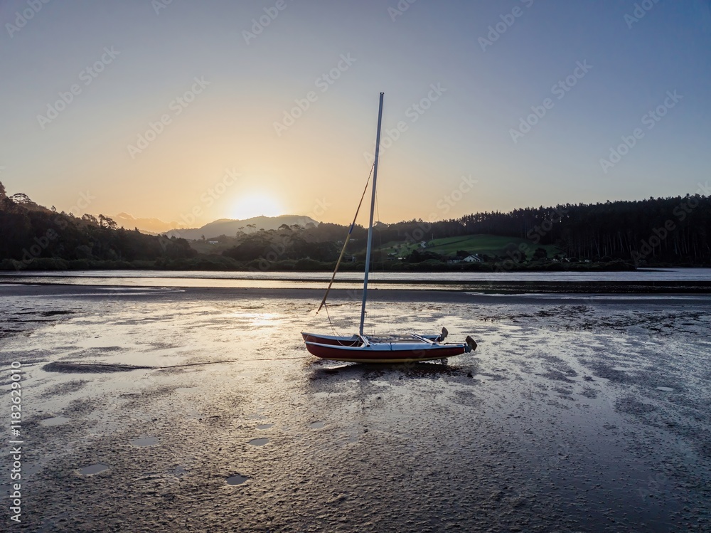 Sunrise over a tranquil bay; a small catamaran rests on the mudflats. Peaceful morning scene. , Point Wells, Auckland, New Zealand Sunrise over a tranquil bay; a small catamaran rests on the mudflats. Peaceful morning scene. , Point Wells, Auckland, New Zealand
