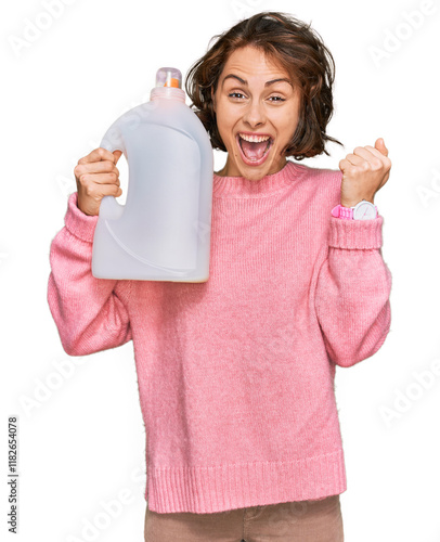 Young hispanic woman doing laundry holding detergent bottle screaming proud, celebrating victory and success very excited with raised arms