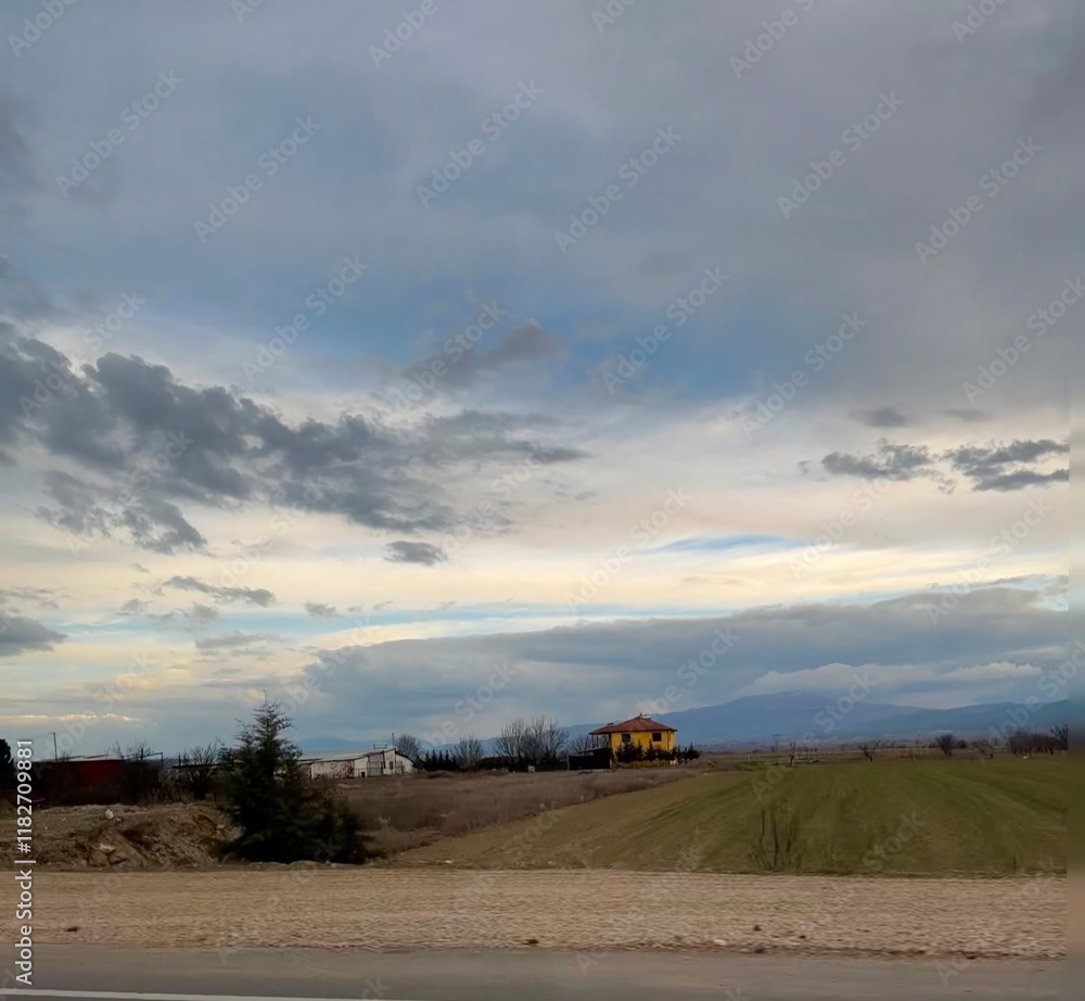 Fototapeta premium Scenic countryside landscape with a dramatic cloudy sky, a distant yellow house, and green fields. Serene rural view with natural colors and open space
