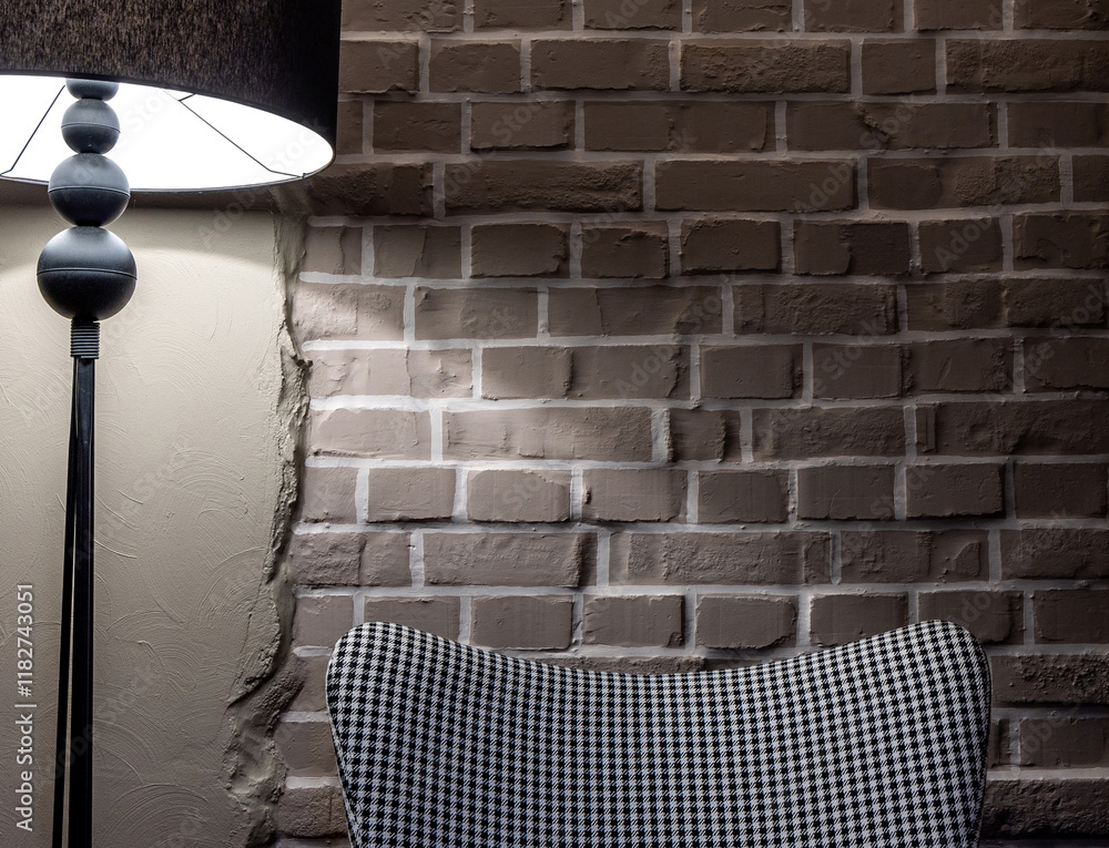 A grey, comfortable armchair with a houndstooth pattern stands against a grey brick wall. The scene is lit by the atmospheric light of a floor lamp.