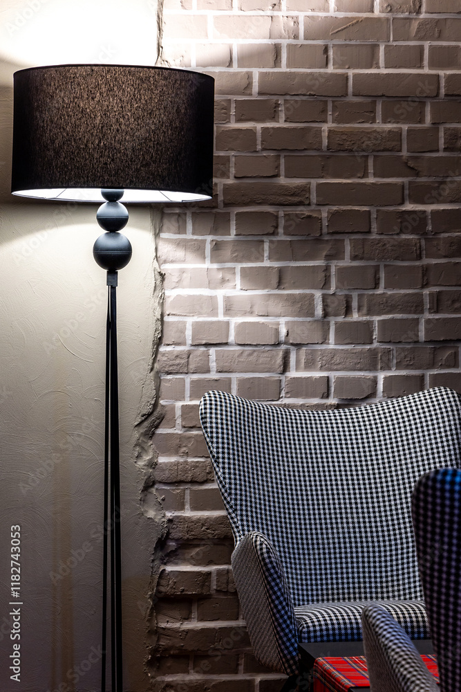 A grey, comfortable armchair with a houndstooth pattern stands against a grey brick wall. The scene is lit by the atmospheric light of a floor lamp.