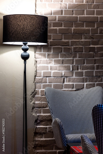 Obraz na płótnie A grey, comfortable armchair with a houndstooth pattern stands against a grey brick wall. The scene is lit by the atmospheric light of a floor lamp. w salonie