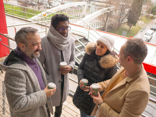 A group of four business colleagues taking a coffee break outdoors on a winter day. They are standing together, smiling, and holding coffee cups in an urban business district. Dressed in warm coats