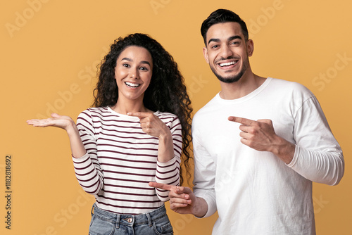 Check This. Cheerful Middle Eastern Couple Pointing At Open Palm, Happy Arab Man And Woman Demonstrating Invisible Object, Showing Copy Space While Standing Isolated On Yellow Background, Panorama photo