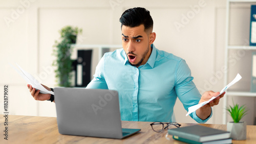 Shocked young Arab businessman with documents looking at laptop screen in dismay, shouting WTF, making mistake in project at office. Business crisis, error, deadline concept
