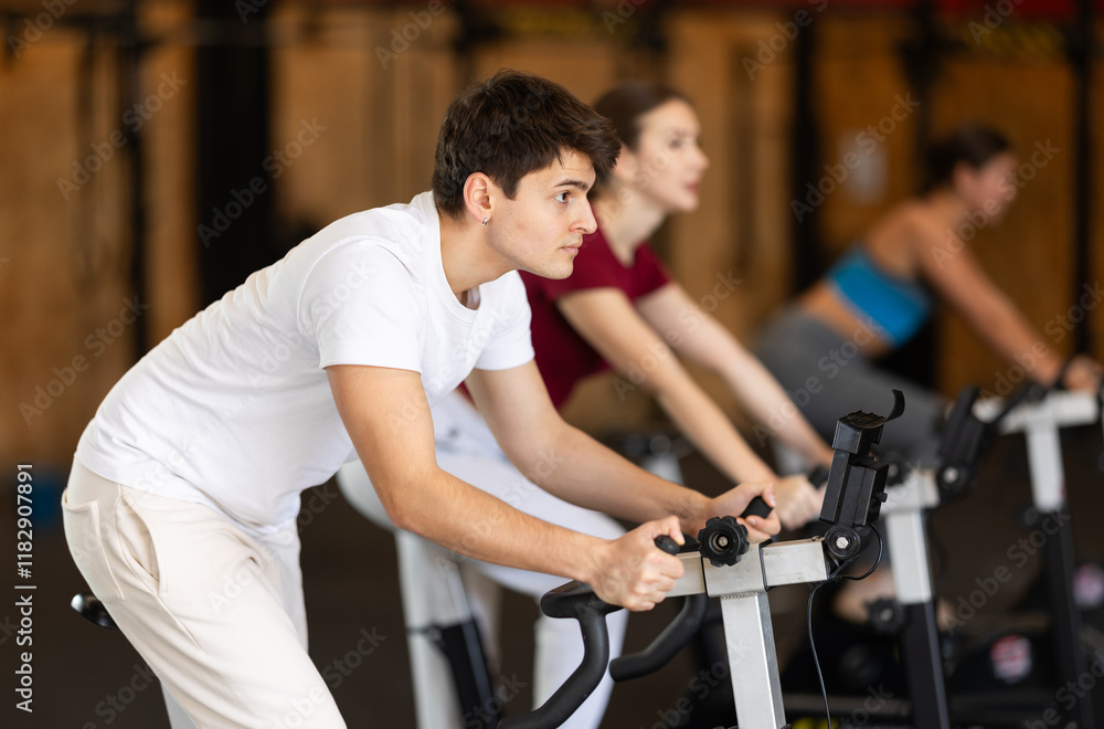 Athletic man at group cycling training in the gym. Guy is actively pedaling on an exercise bike against the background of training people Athletic man at group cycling training in the gym. Guy is actively pedaling on an exercise bike against the background of training people