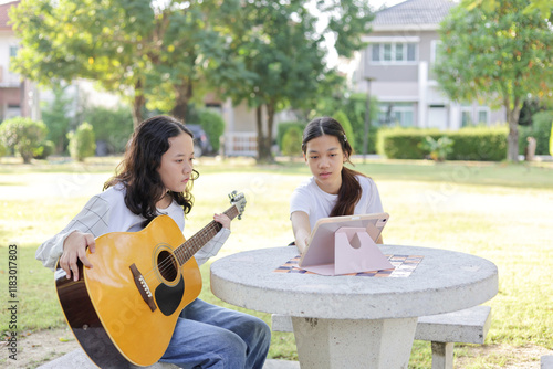 Asian kid girl and friend learning online guitar lesson in the park, Friends playing guitar on picnic in park