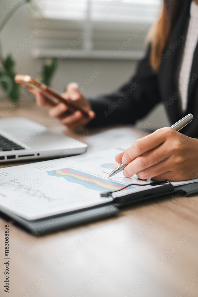 Fototapeta premium Professional woman reviewing charts and graphs at a desk, using a tablet and smartphone for business analysis.