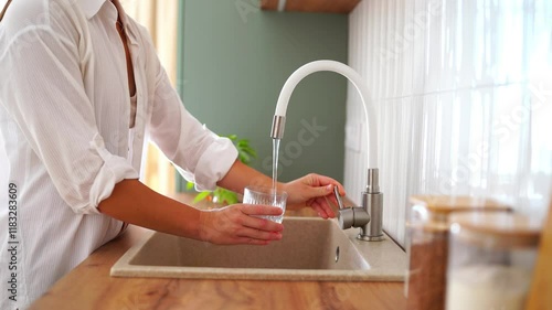Woman fills a glass with clean filtered tap water for drinking in scandinavian style kitchen at home