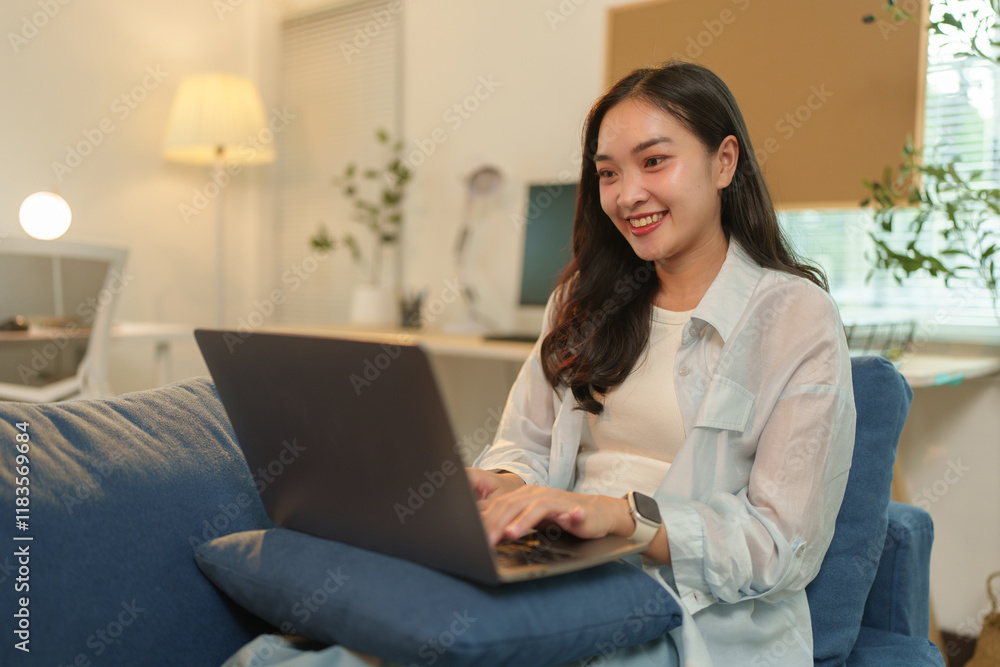 Smiling young asian businesswoman is working remotely from her home office, comfortably sitting on a sofa and using a laptop, enjoying the convenience and flexibility of working from home Smiling young asian businesswoman is working remotely from her home office, comfortably sitting on a sofa and using a laptop, enjoying the convenience and flexibility of working from home