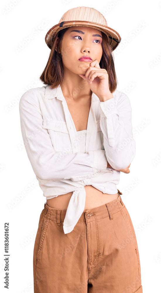 Young beautiful chinese girl wearing explorer hat serious face thinking about question with hand on chin, thoughtful about confusing idea Young beautiful chinese girl wearing explorer hat serious face thinking about question with hand on chin, thoughtful about confusing idea