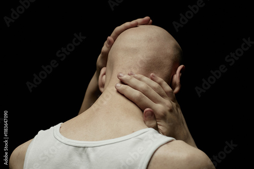 Rear view of pale bald man touching skin of neck and head in feeling of deep regret or anxiety while standing in studio with low key light, copy space