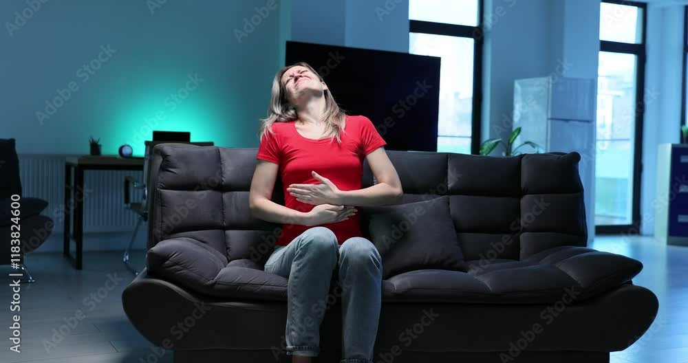 Young woman experiences intense abdominal pain sitting on sofa in modern living room. Female touches stomach suffering from menstrual cramps