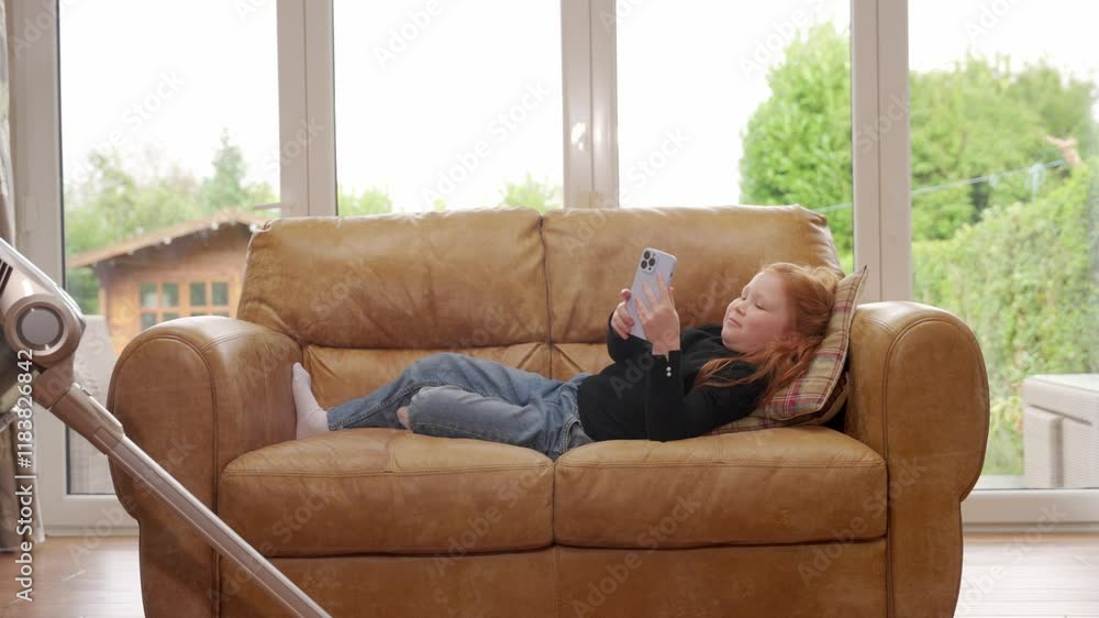 Young girl relaxing on couch and using smartphone while mother vacuuming room Young girl relaxing on couch and using smartphone while mother vacuuming room