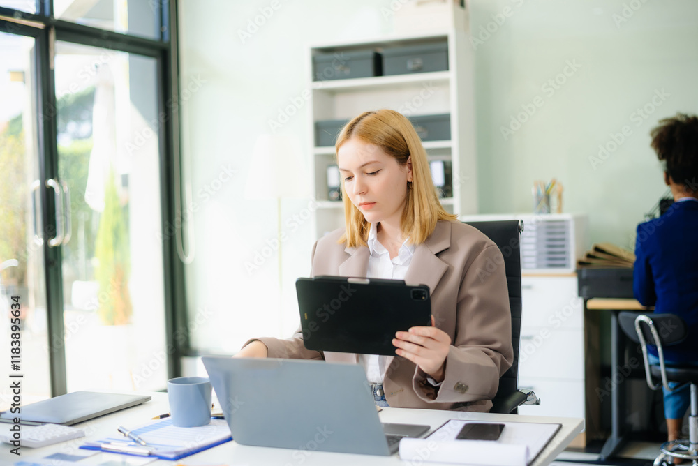 Obraz premium Young woman, smiling and working at her desk in an office environment. She is wearing a business suit