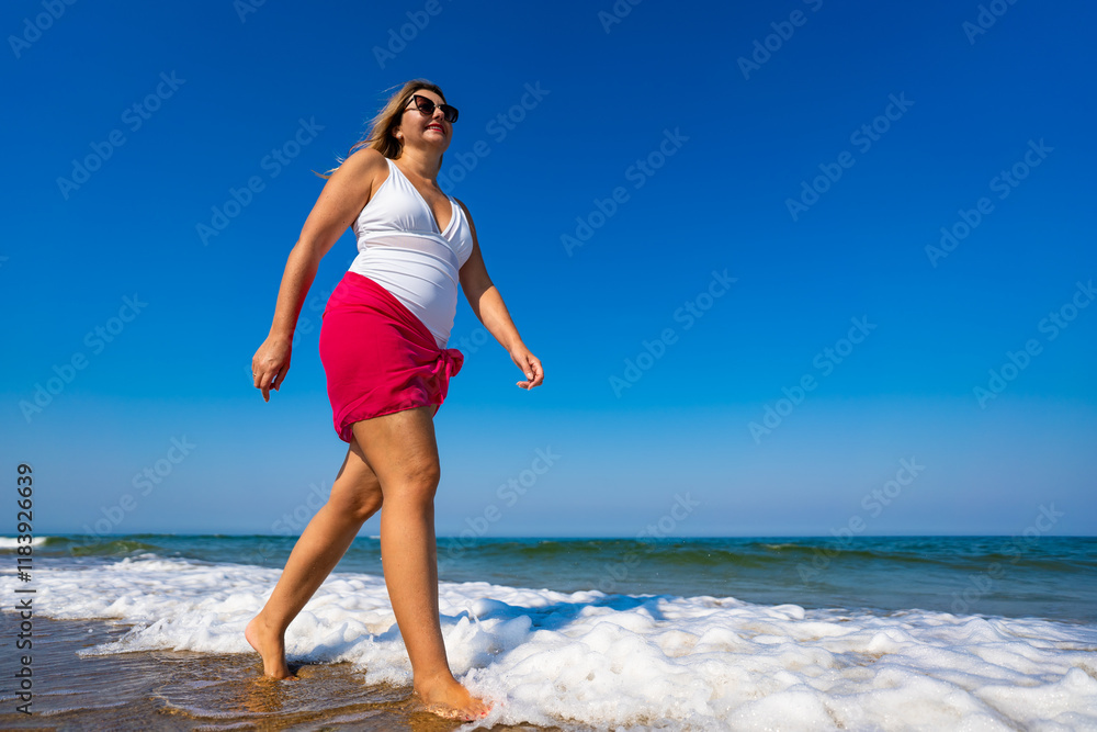 Beautiful middle-aged woman walking on sandy beach in summertime. Physical activity on beach. Side view Beautiful middle-aged woman walking on sandy beach in summertime. Physical activity on beach. Side view