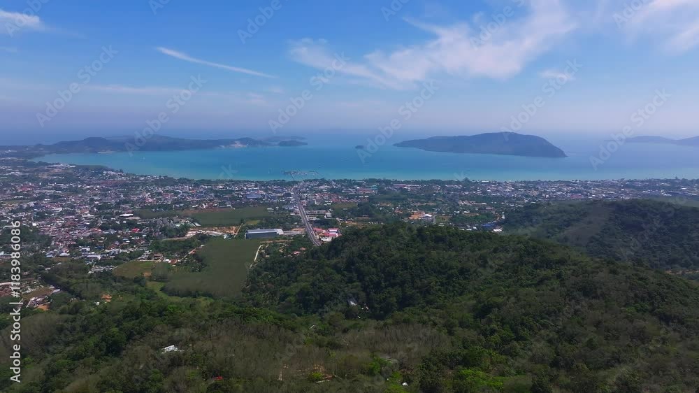 Aerial perspective of Phuket Island, showing green hills, urban areas, dense forests, and the turquoise coastline meeting the Andaman Sea. Aerial perspective of Phuket Island, showing green hills, urban areas, dense forests, and the turquoise coastline meeting the Andaman Sea.