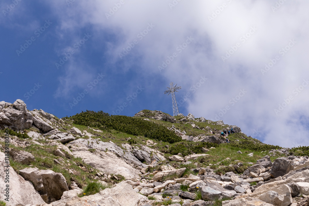 Fototapeta premium A part of the mountain slope with bushes and stones, and a road leading to the top of the mountain, where there is a large cross against the sky with white clouds.
