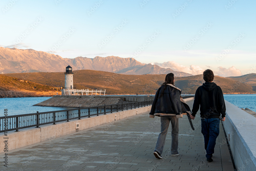 Fototapeta premium A couple walks hand in hand along a scenic coastal promenade, heading towards a lighthouse at sunset. The picturesque view features calm blue waters, rugged hills, and a warm golden light