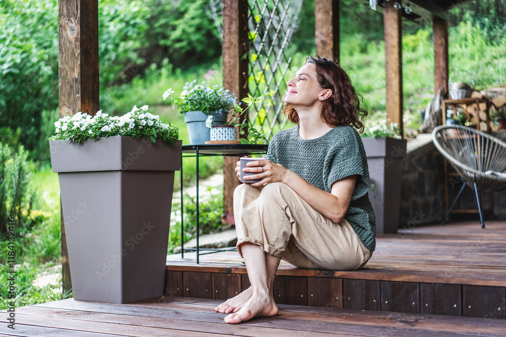 Young happy smiling Caucasian woman sitting on the wooden terrace of a country house enjoying the summer weather
