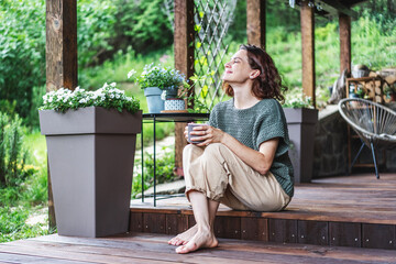 Young happy smiling Caucasian woman sitting on the wooden terrace of a country house enjoying the summer weather
