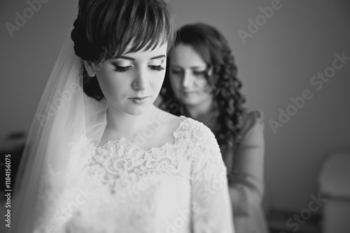 A woman is getting ready for her wedding and her mother is helping her. The bride is wearing a white dress with lace detailing and a veil. The mother is adjusting the bride's dress
