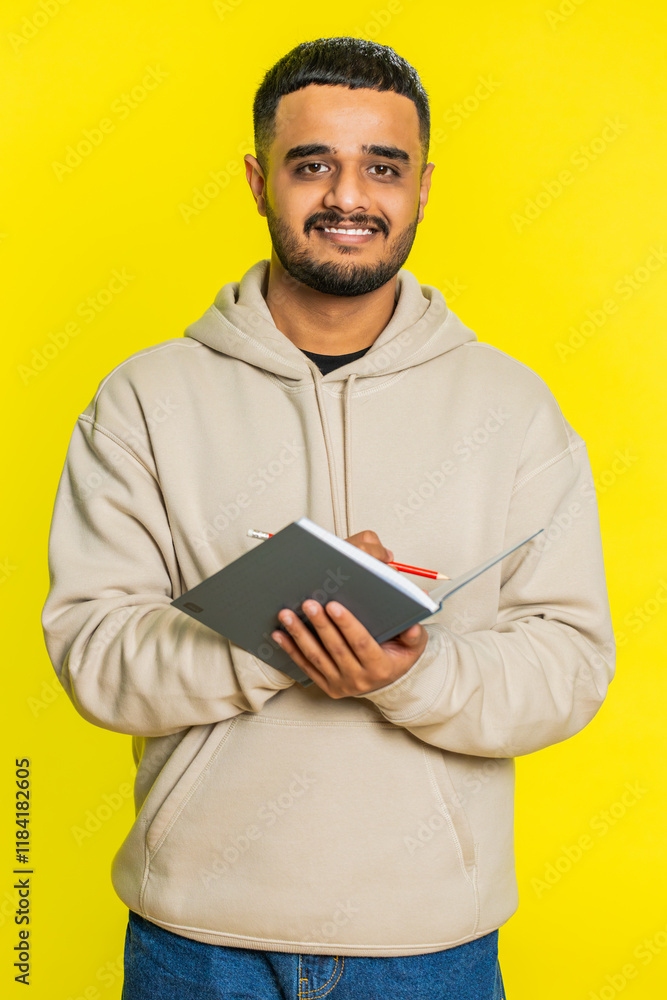 Thoughtful journalist young Indian man making notes, writing down thoughts with pen into notepad notebook diary, to do list, good idea inspiration. Arabian guy isolated on yellow background. Vertical Thoughtful journalist young Indian man making notes, writing down thoughts with pen into notepad notebook diary, to do list, good idea inspiration. Arabian guy isolated on yellow background. Vertical