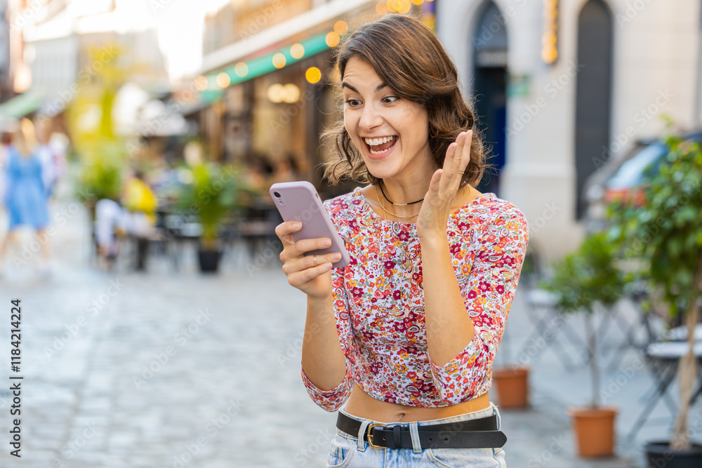 Fototapeta premium Young woman use mobile smartphone celebrating win good message news, lottery jackpot victory, giveaway online, play game outdoors. Happy girl tourist standing in urban city street. Town lifestyles