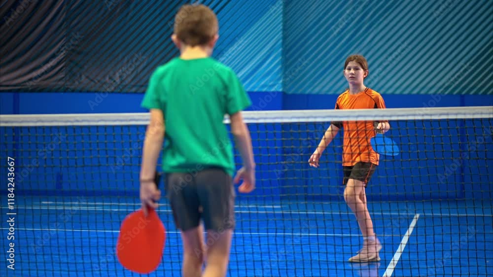 A boy and a girl playing pickleball on a blue, inside court A boy and a girl playing pickleball on a blue, inside court