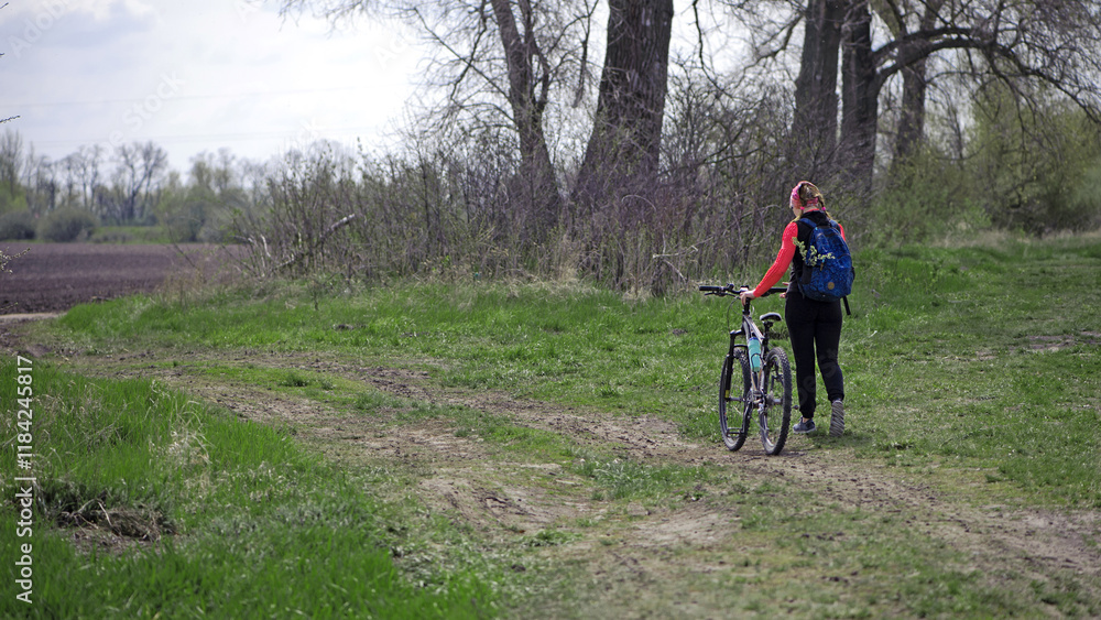 Fototapeta premium a girl with a bicycle walks a field trail. Woman riding a mountain bike in the forest. Hiker woman with backpack. one young woman riding a bike in nature. rolling the bike in his hands. back view.