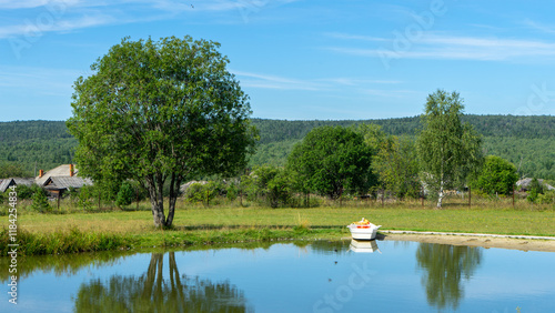 Small non-motorized or motorless white pleasure boat on shore of pond or lake in village or rural area. Rural pastoral landscape. Outdoor recreation in good environment photo