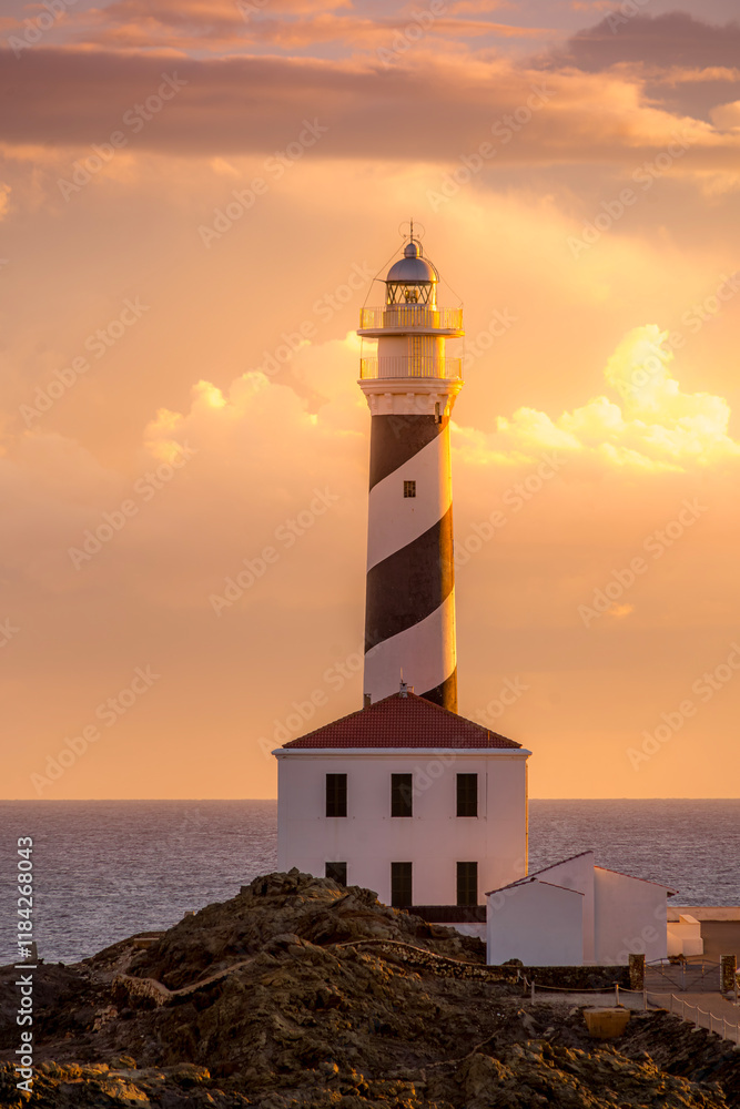 Favaritx Lighthouse at dawn, located on the island of Menorca in the Balearic Islands, Spain, offering serene coastal views and a striking contrast of light against rocky cliffs