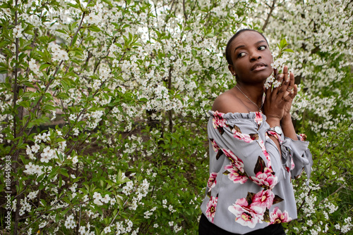 African american woman gently caressing white blossoming tree branches, breathing floral fragrance during springtime in peaceful orchard landscape. Meditation, mental treatment, psychology