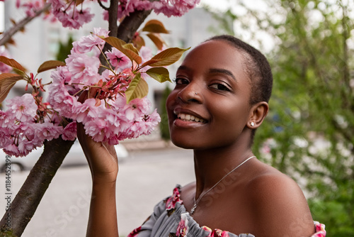 Young black woman smelling cherry blossoms in spring wearing floral top, enjoying the scent of sakura flowers in springtime, with pink flowers and green leaves in background. Japanese Sakura Flower