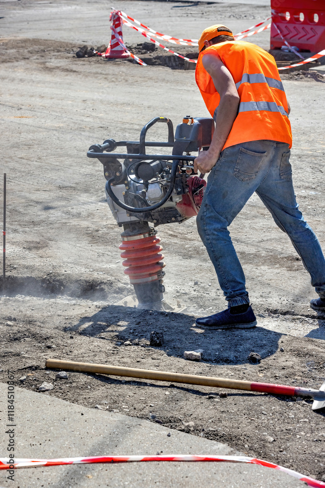 Construction worker uses a vibratory tamper to compact soil at a site under bright sunlight