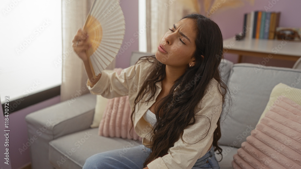 Young woman fanning herself in a cozy living room with pastel decor and comfortable seating Young woman fanning herself in a cozy living room with pastel decor and comfortable seating