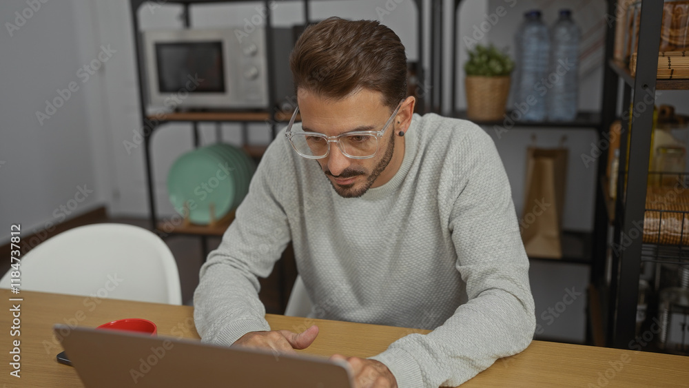 Obraz premium Young man with glasses focused on his laptop in an office setting, surrounded by kitchenware and a microwave, indicating a modern workspace.