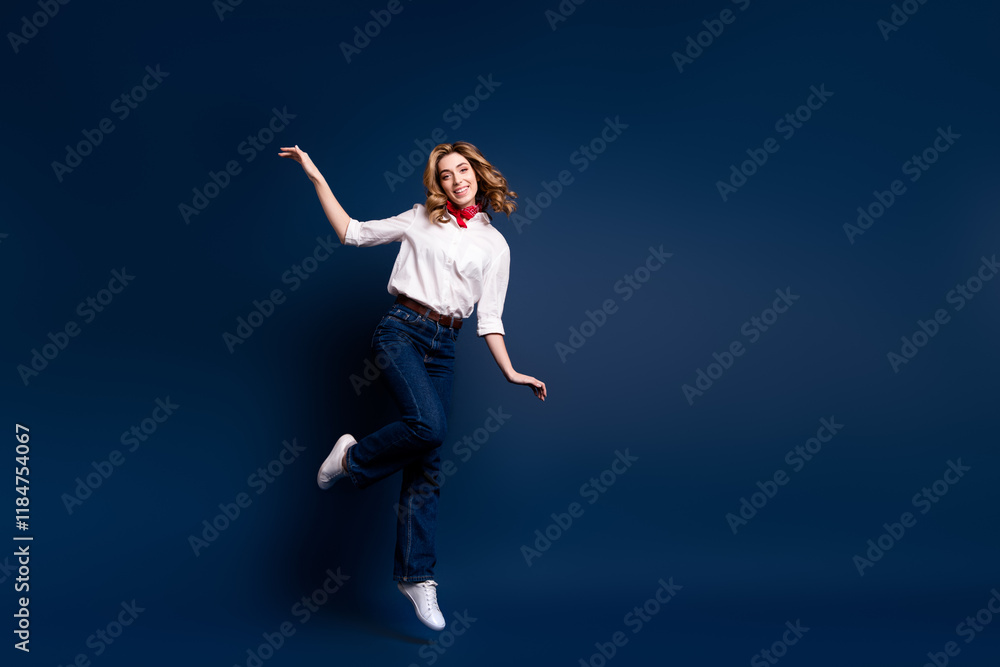 Obraz premium Joyful young woman in white shirt and jeans jumping with elegance on dark blue background