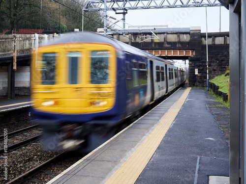 Blurred train speeds through a railway station under an overpass on a rainy day.