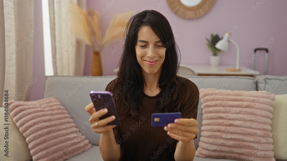Woman smiling in living room holding smartphone and credit card, engaging in online shopping while sitting on a cozy sofa with pastel cushions Woman smiling in living room holding smartphone and credit card, engaging in online shopping while sitting on a cozy sofa with pastel cushions