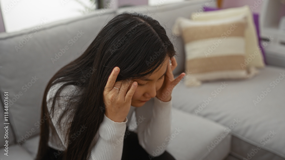 Woman sitting indoors looking stressed with hands on temples in living room showcasing a cozy home environment with a comfortable sofa and soft cushions nearby. Woman sitting indoors looking stressed with hands on temples in living room showcasing a cozy home environment with a comfortable sofa and soft cushions nearby.