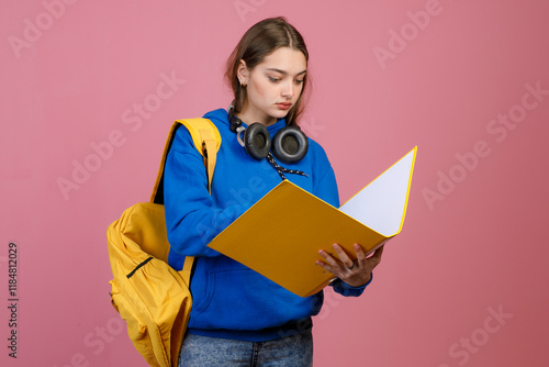 Front view of brunette schoolgirl standing, holding open folder, reading. Pretty young female wearing blue khudi and jeans, earphones, looking down. Concept of modern lifestyle. photo