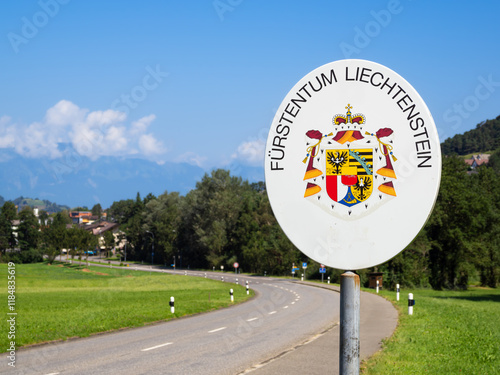 A road sign indicating the entrance to the Principality of Liechtenstein, featuring the coat of arms. The background shows a clear blue sky and green fields.
