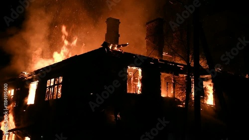 House is on fire at night, flames inside at home, burning window. The wooden building is ablaze, completely engulfed in fire. Loss of housing, disaster, trouble for unhappy people.