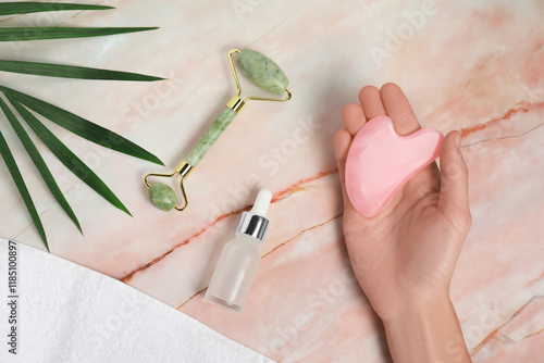 Woman with gua sha tool, face roller, cosmetic product, towel and leaf on pink marble background, top view photo